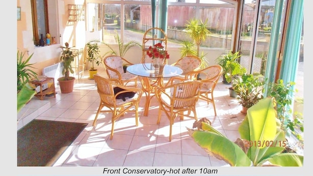 A conservatory with a round table and wicker chairs, surrounded by potted plants and a view of the street. - Inviting 6-Bed Farmhouse (Cravans)