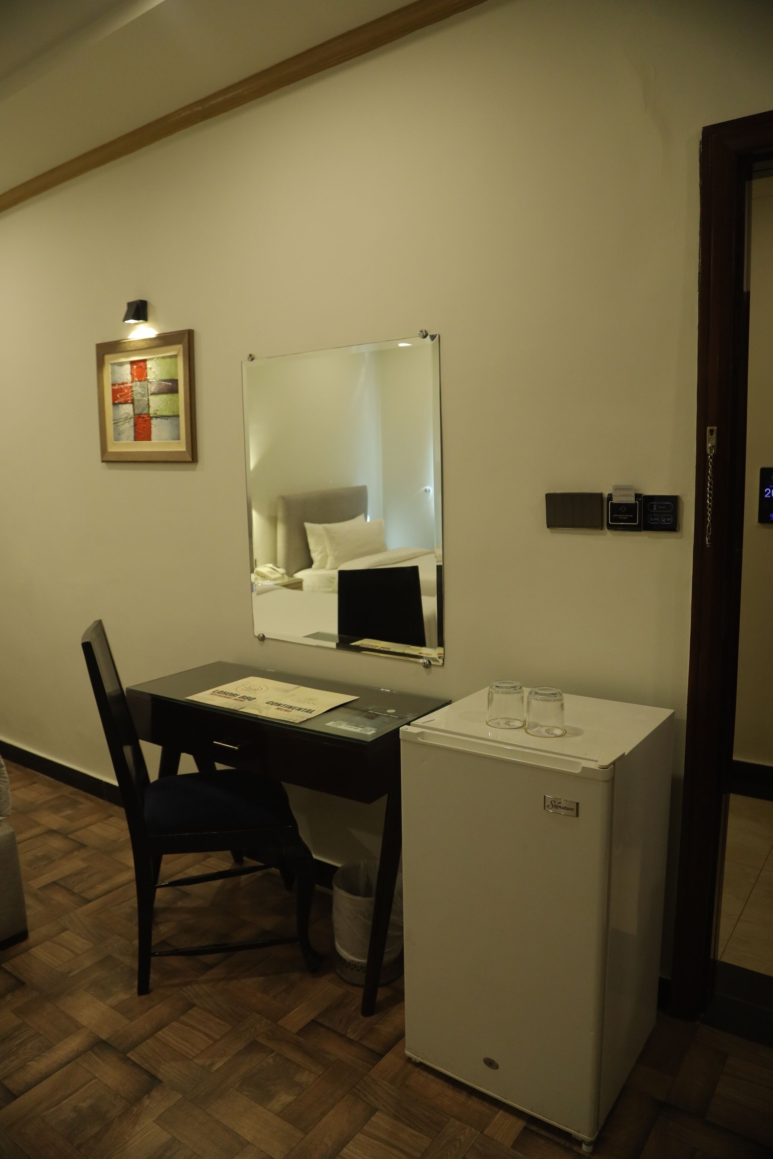 A compact hotel room with a wooden floor, a desk with a chair, a small white refrigerator, a mirror, and a bedside table.