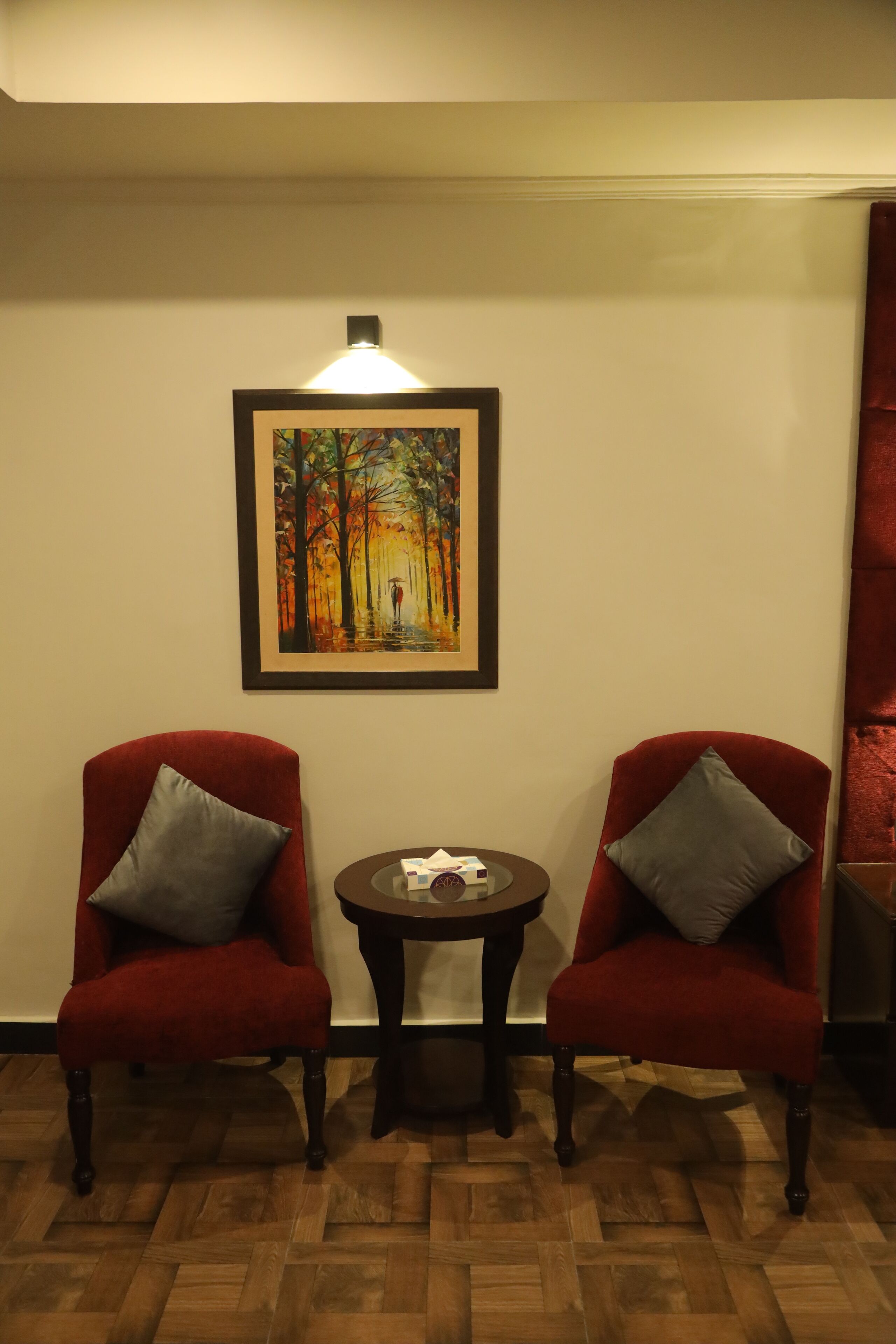 Dos sillones rojos con cojines grises, una mesita redonda con un control remoto y un cuadro enmarcado que representa un paisaje forestal en la pared.