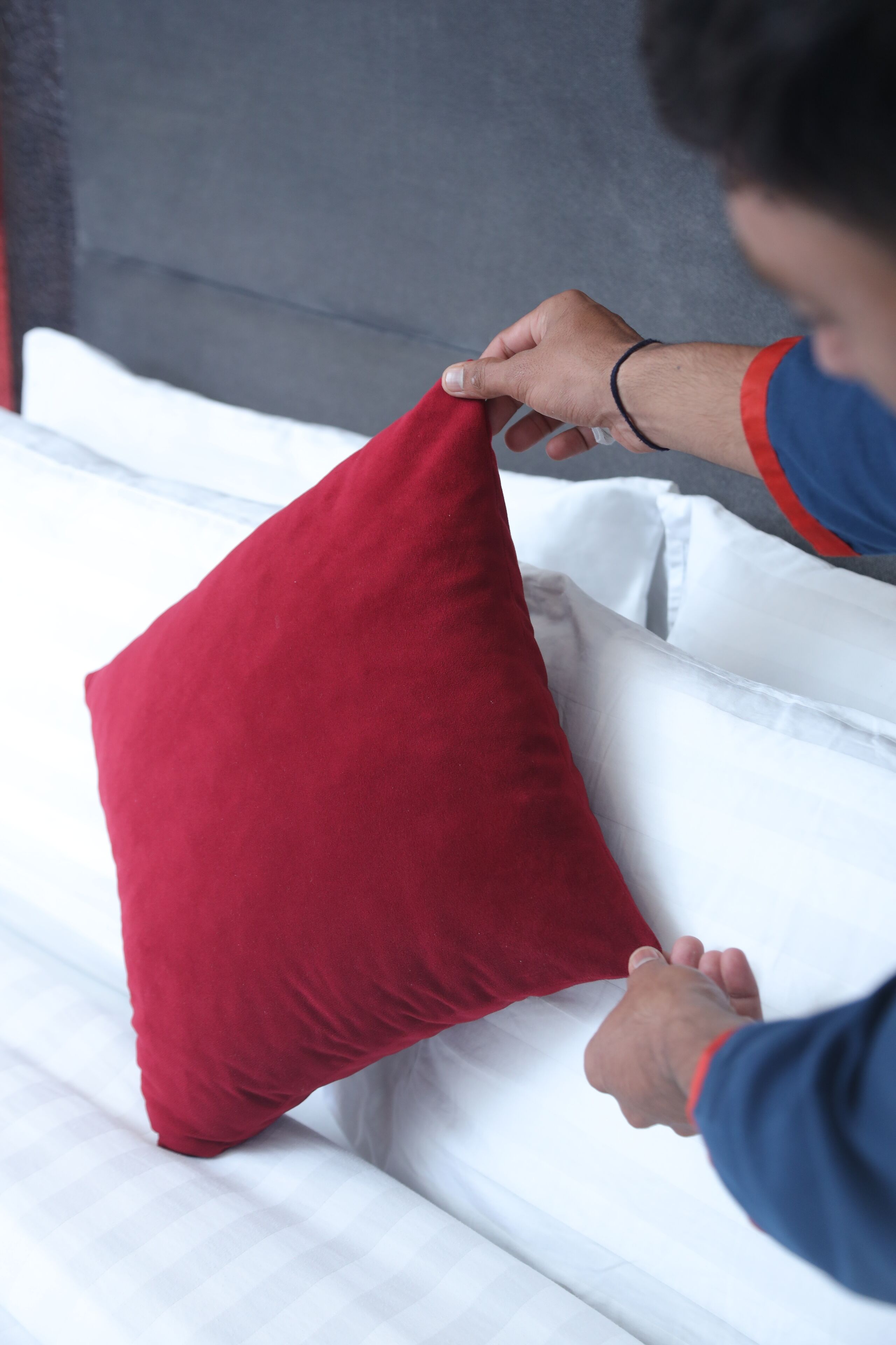 A person adjusting a red pillow on a white bed with a grey headboard.