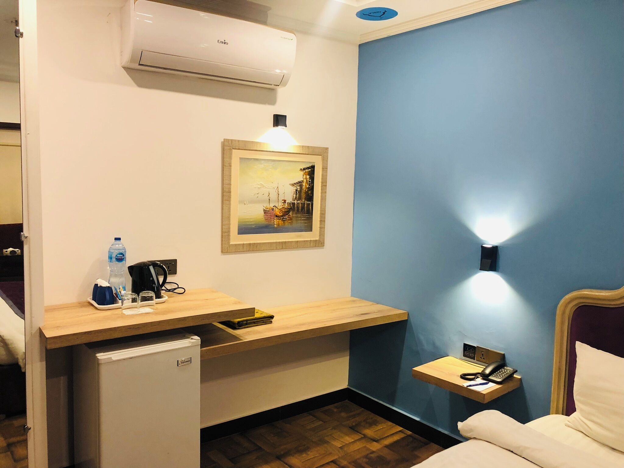 A hotel room with a desk, a small refrigerator, a painting, a bedside table with a lamp, and a bed.