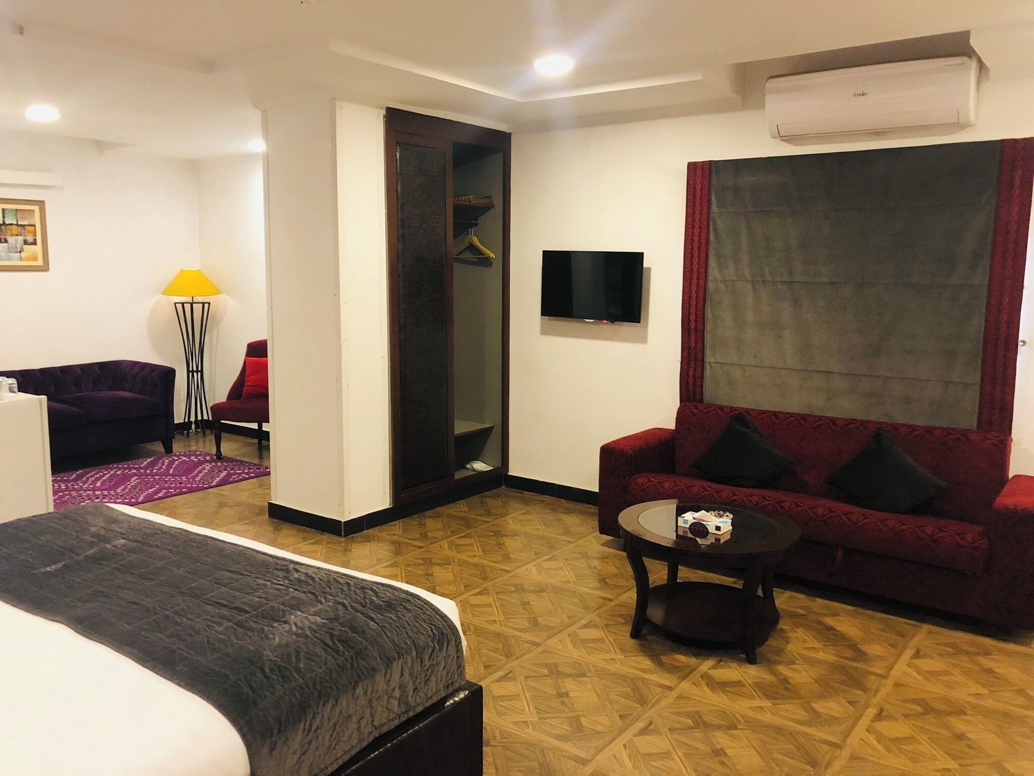 A hotel room with a bed, a red sofa, a small table with a dog, a TV, and an air conditioning unit.