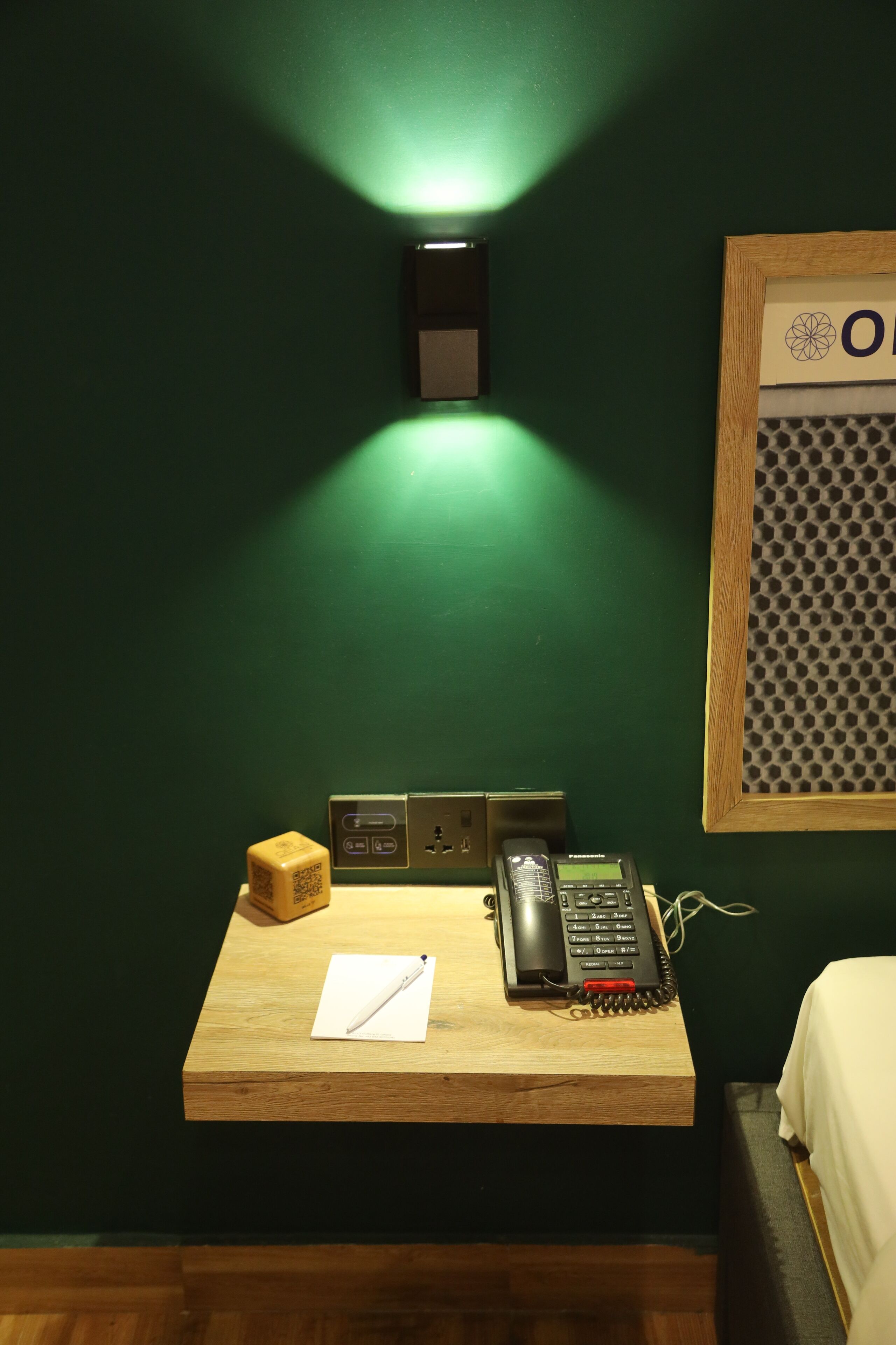 Une chambre d’hôtel avec un bureau en bois, un téléphone, un bloc-notes, une petite boîte et un luminaire fixé au mur.