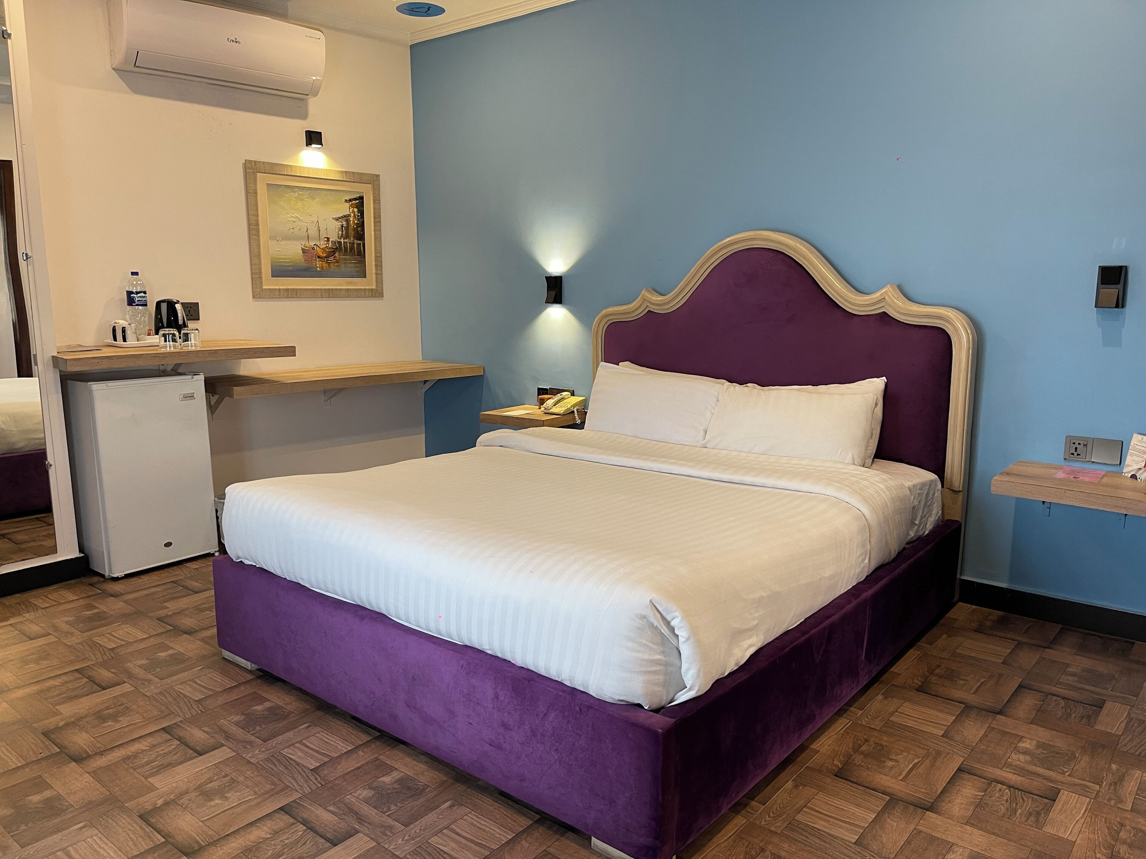 A hotel room with a purple bed, a small desk with a lamp, and a painting on the wall.