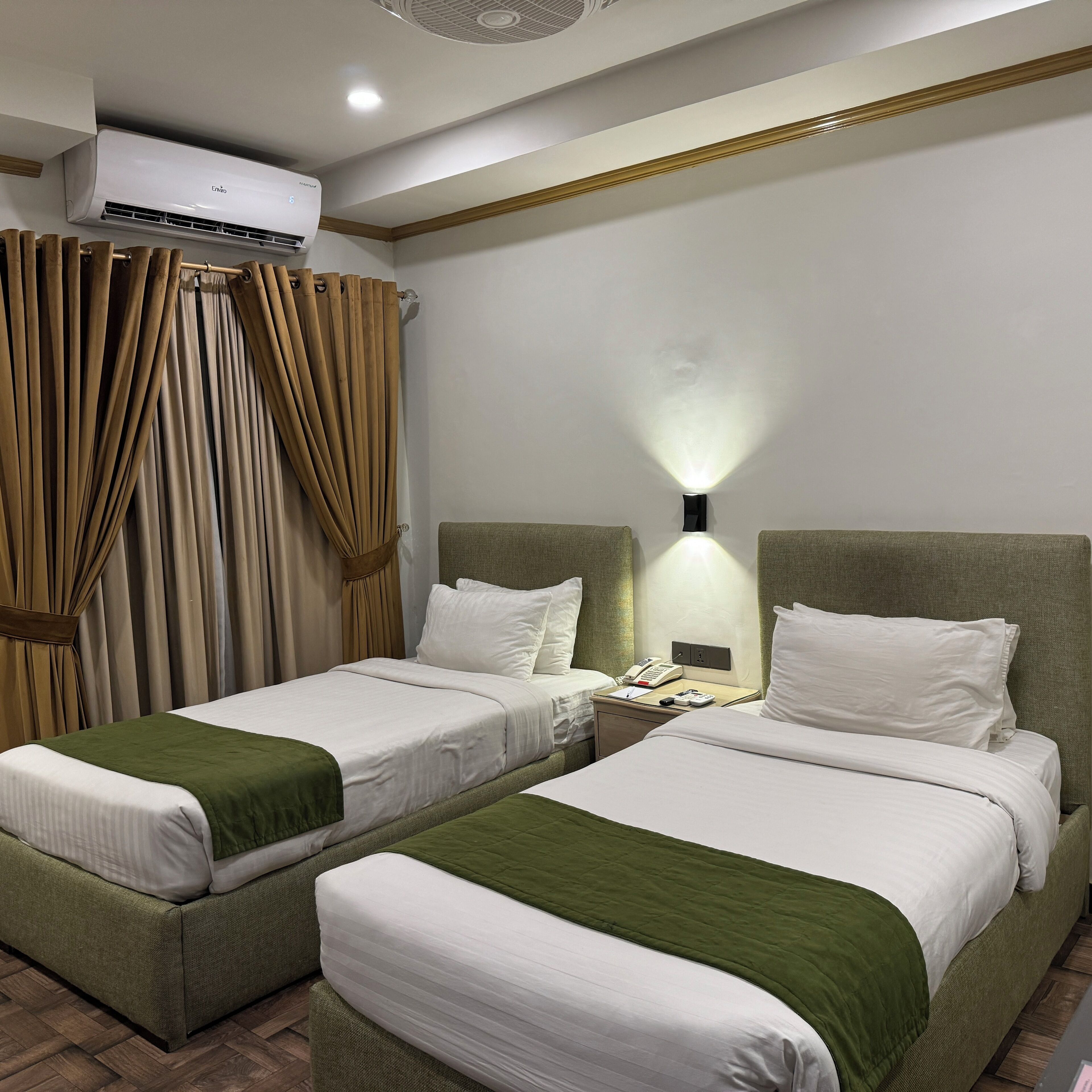 A hotel room with two beds, a wall-mounted air conditioner, a bedside lamp, and a telephone.
