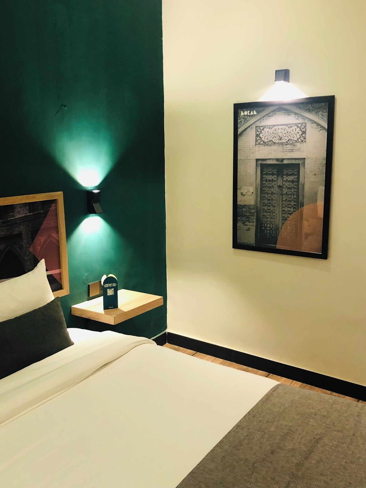 A hotel room with a bed, a nightstand with a lamp, a framed picture on the wall, and a small table with a QR code.