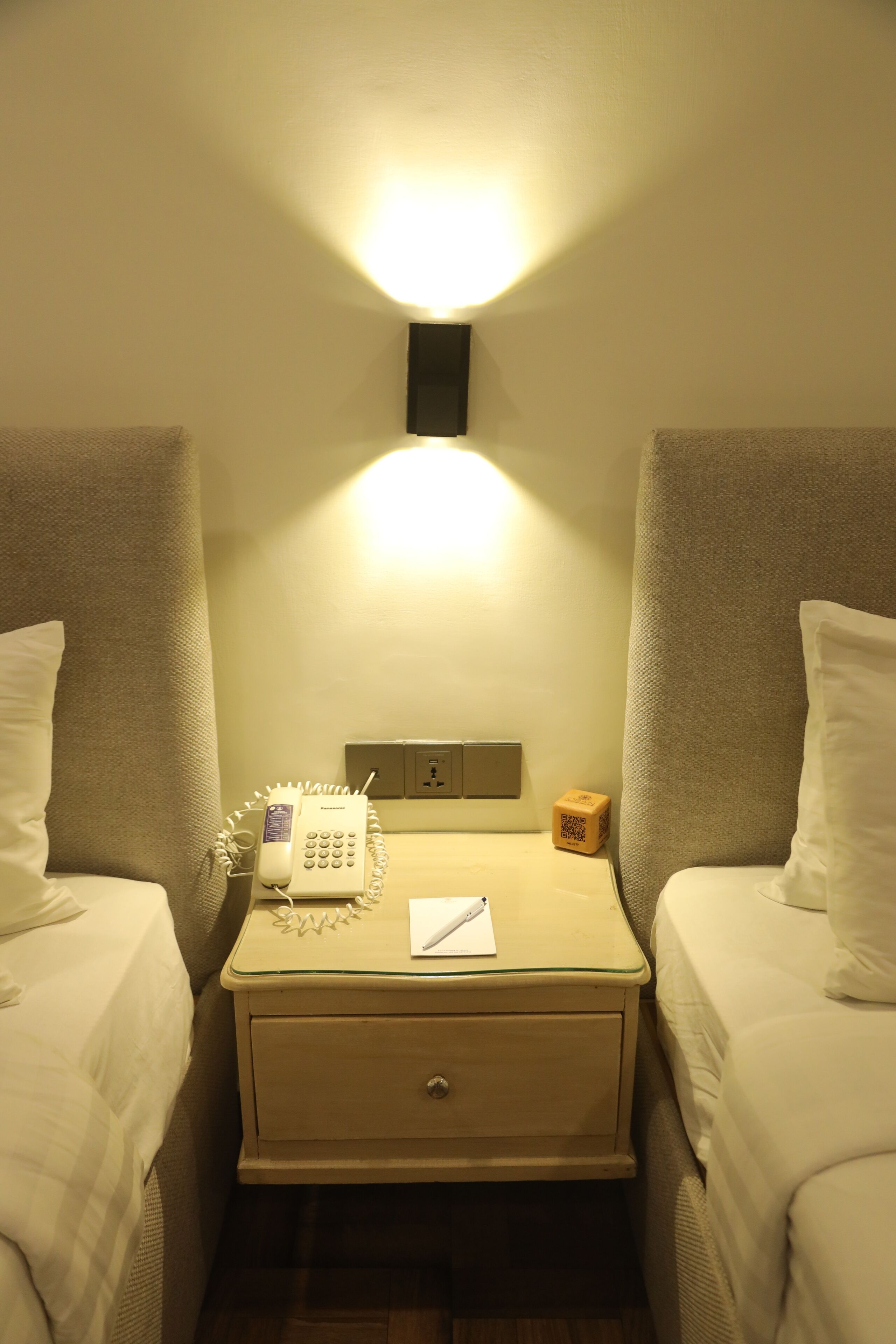 A hotel room with two beds, a bedside table with a phone, a notepad, and a QR code.