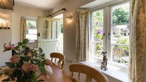 A dining area with a wooden table, chairs, and a vase of flowers. There is a window with curtains and a view of the outside. - Stunning 2 Bed Cottage in Fordwells (Fordwells)