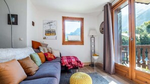 A cozy living room with a sofa, colorful cushions, a knitted pouf, a window with a view of mountains, and a balcony with a railing.