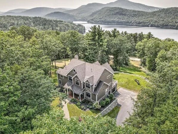 A large, two-story house with a prominent front porch, surrounded by dense forest and a lake in the background.