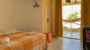 A hotel room with a bed, a small orange ottoman, a table with a colorful pattern, and a sliding glass door leading to a balcony. - Pousada A Praia (Parnamirim)