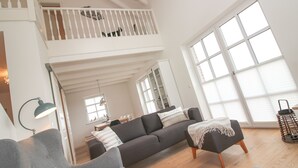 Ein geräumiger Wohnbereich mit einem grauen Sofa, einem kleinen Hocker und einer Treppe, die auf einen Balkon führt.