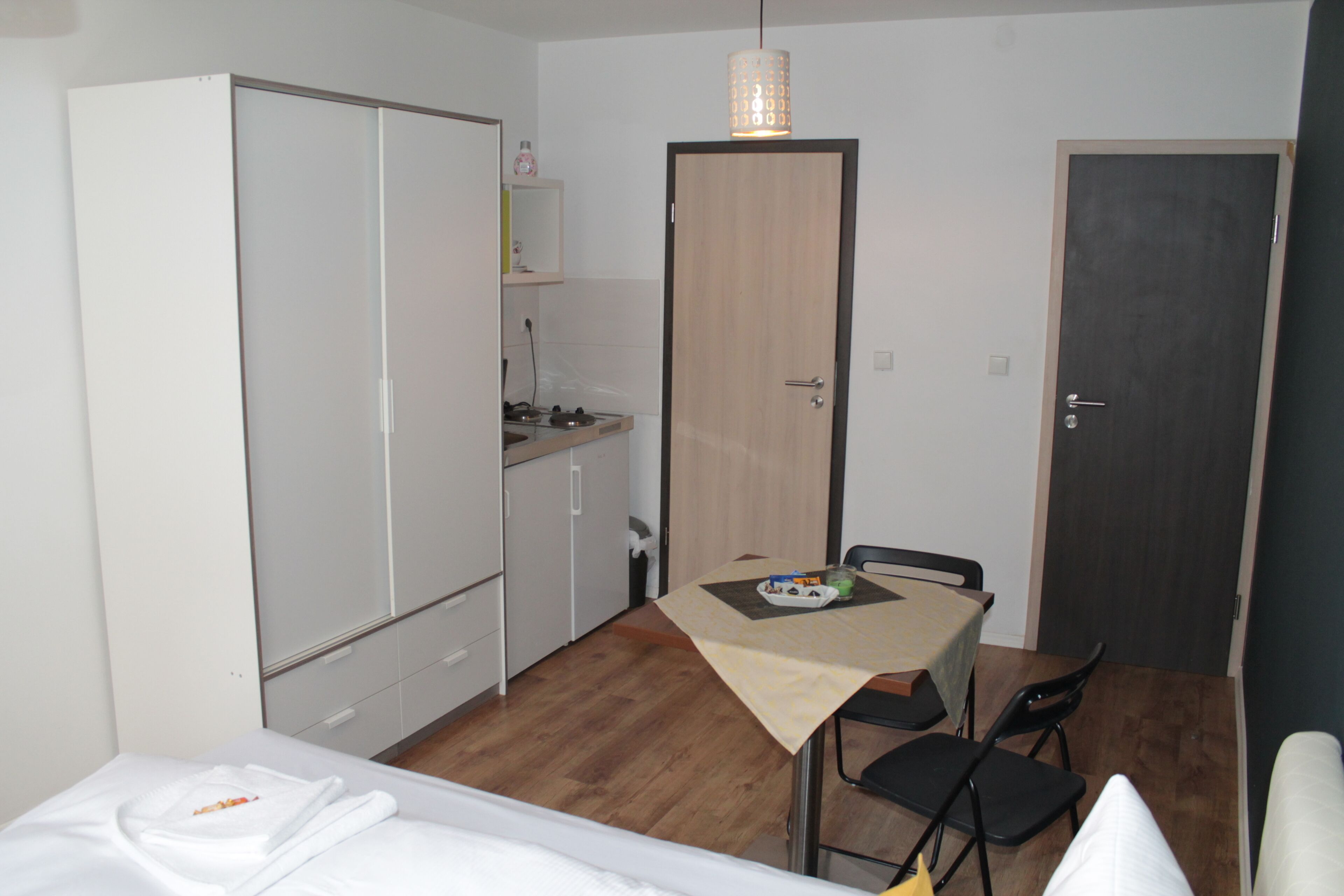 Quarto de hotel pequeno, com cama, uma kitchenette, uma mesa de jantar e cadeiras, e um guarda-roupa.