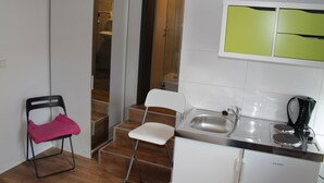 A compact kitchen with a sink, a coffee maker, and a refrigerator. There are two chairs, one with a pink cushion and another plain white. A wooden staircase leads to a bathroom. - Pension Michelangelo (Eisenach)