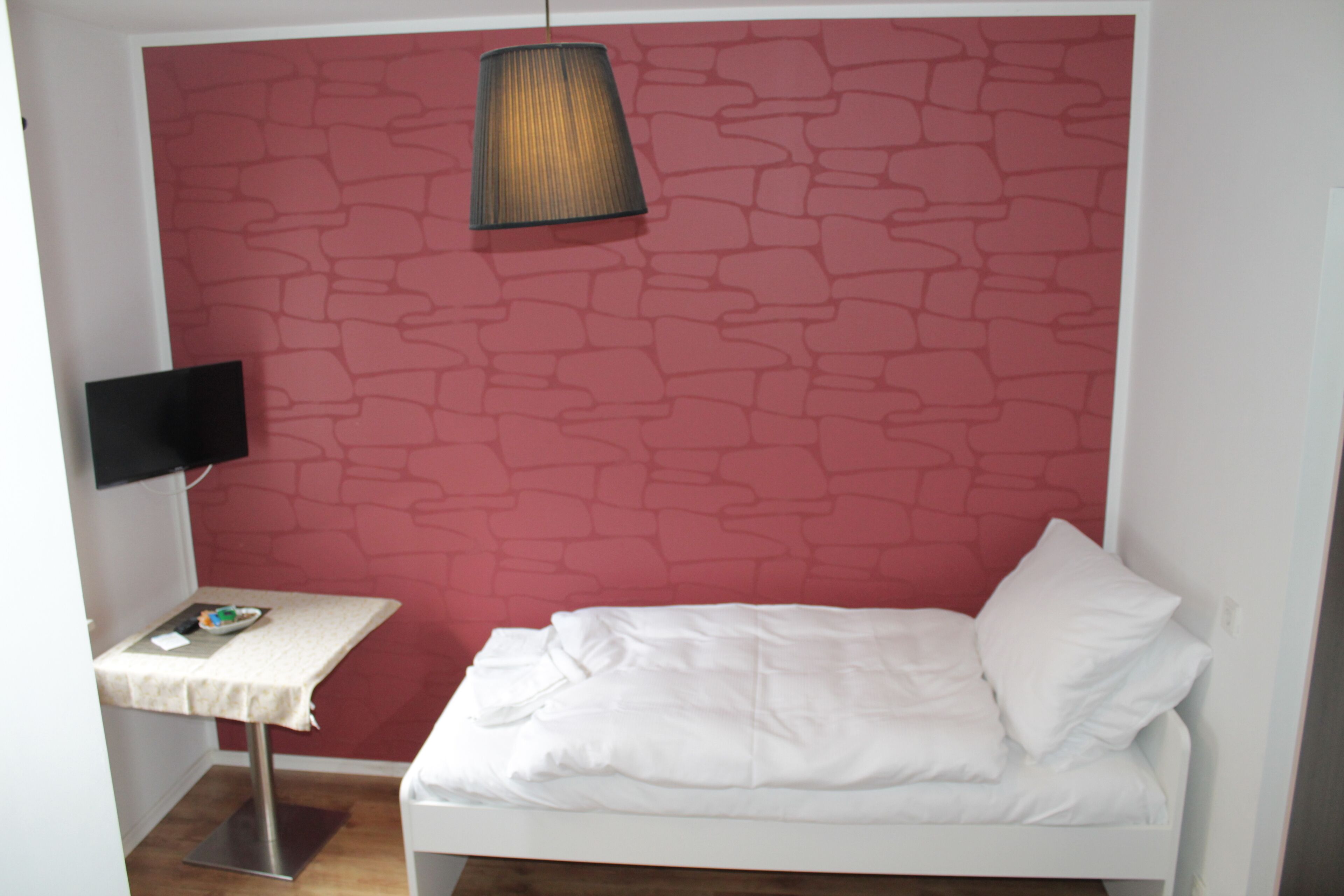 A small room with a white bed, a table with a lamp, and a TV on a stand.