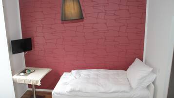 A small room with a white bed, a table with a lamp, and a TV on a stand.