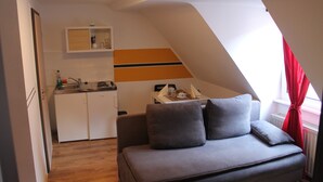 A compact living space with a kitchenette, a grey sofa, and a window with red curtains. - Pension Michelangelo (Eisenach)