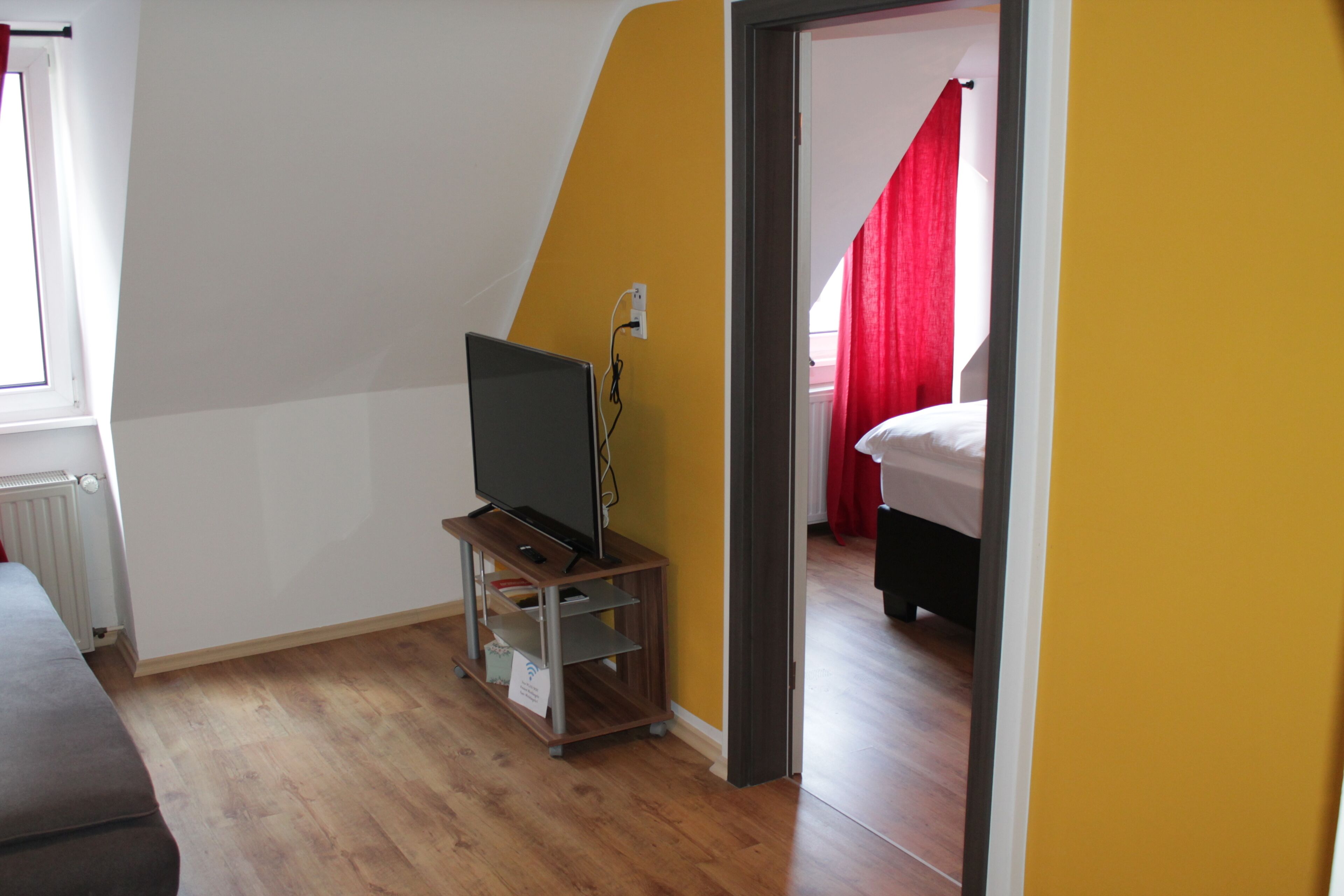 A room with a television on a wooden stand, a radiator, and a bed with red curtains.