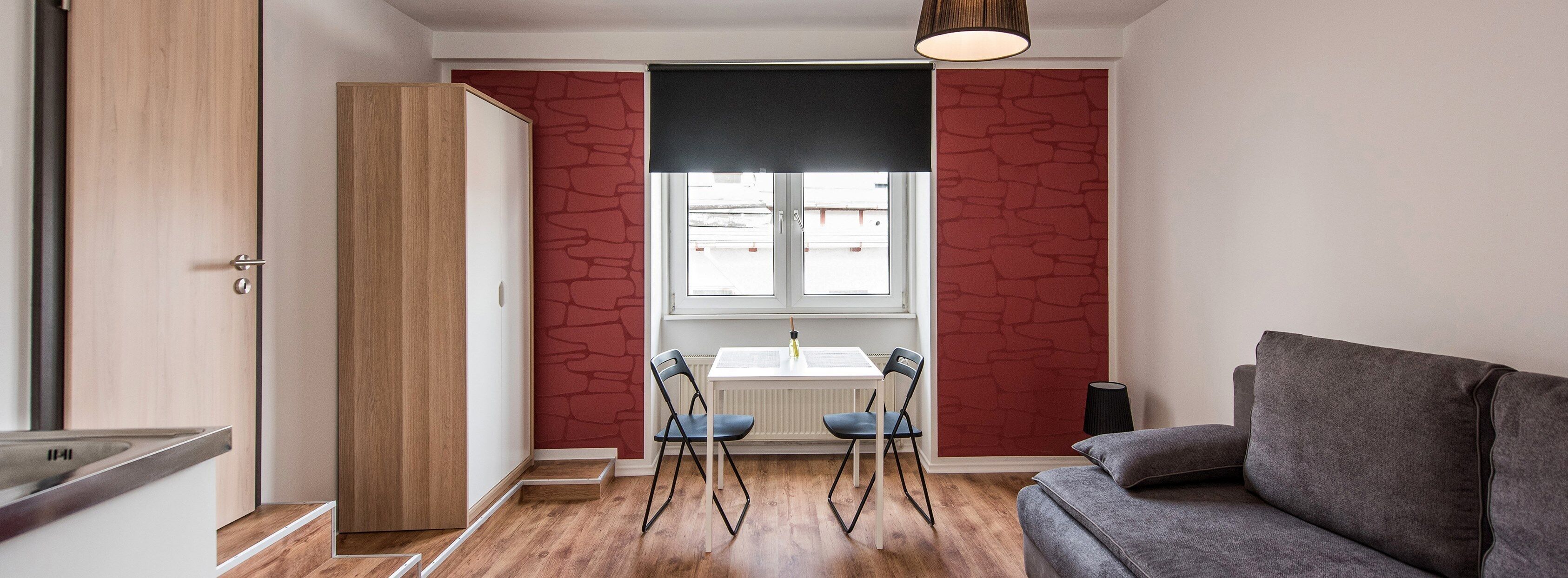 Ett modernt vardagsrum med en grå soffa, en trägarderob, ett matbord med två stolar och ett fönster med en vägg av rött tegel.