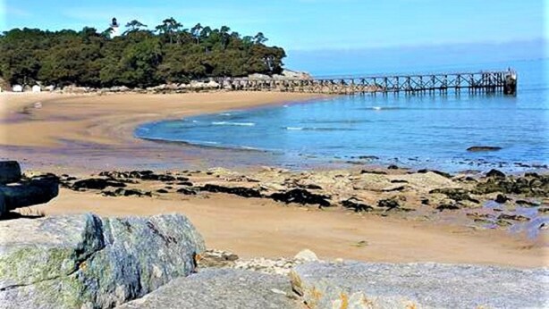 Una playa con un muelle de madera que se adentra en aguas tranquilas, rodeada de rocas y vegetación escasa.