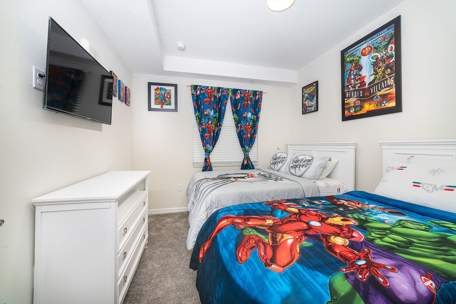 A bedroom with a bed, a dresser, a TV mounted on the wall, and two windows with colorful curtains.