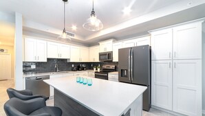 A modern kitchen with white cabinets, a central island, black chairs, and a stainless steel refrigerator.