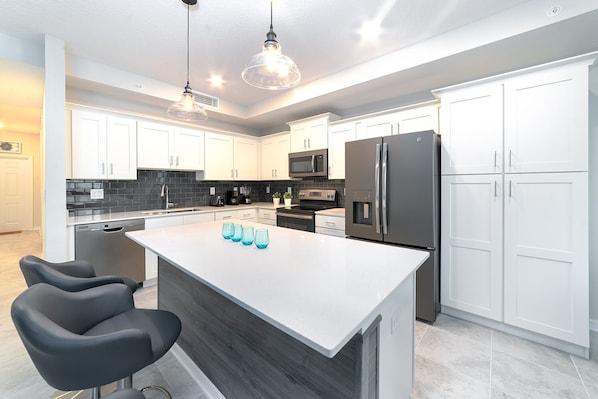 A modern kitchen with white cabinets, a central island, black chairs, and a stainless steel refrigerator.