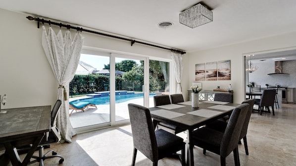 A spacious dining area with a view of an outdoor swimming pool, a kitchen area, and a modern ceiling light fixture.