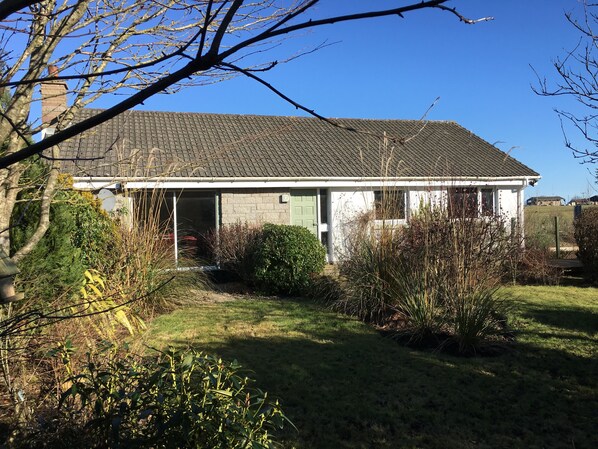Ein eingeschossiges Haus mit Ziegeldach, einer Haustür und einem kleinen Vorgarten mit verschiedenen Pflanzen und Sträuchern.