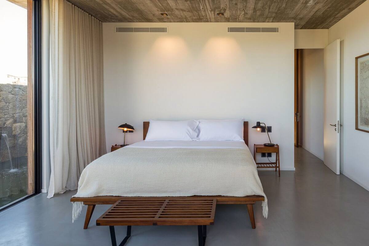 Un dormitorio moderno con una cama grande, lámparas de noche, un banco de madera y un amplio ventanal con cortinas.