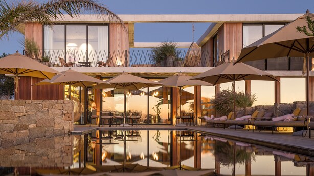 Un área de piscina moderna al aire libre con sillones de descanso y sombrillas, rodeada por un muro de piedra y una terraza de madera.