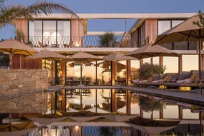 Un área de piscina moderna al aire libre con sillones de descanso y sombrillas, rodeada por un muro de piedra y una terraza de madera.