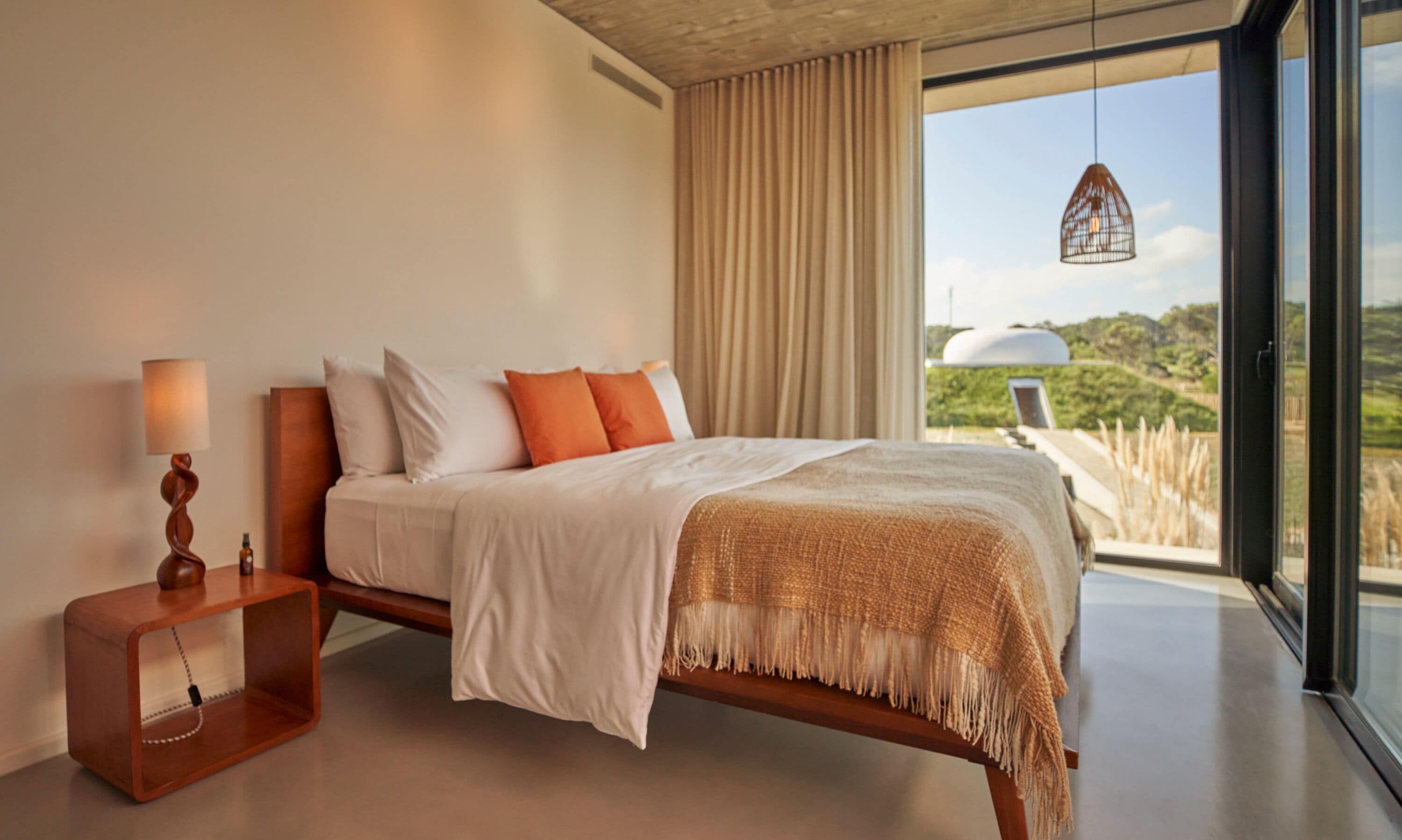 A modern bedroom with a large bed, a wooden bedside table, and a view of the outdoors through large windows.