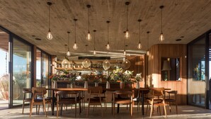 Un interior moderno con techo de madera y lámparas colgantes, una zona de bar con estantes y botellas, y un área de estar con sillas y mesas.