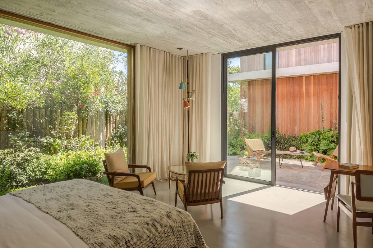 Un dormitorio moderno con una cama grande, un escritorio y una silla. Hay una puerta de vidrio corrediza que da a un balcón con vistas a un jardín.