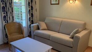 A living room with a beige sofa, a white coffee table, a wicker chair, and a window with patterned curtains. - 3-room cottage near sea and golf in Moliets (Moliets-et-Maa)