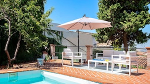 Ein Pool mit gefliestem Deck, weißen Liegestühlen und einem Tisch mit Sonnenschirm.