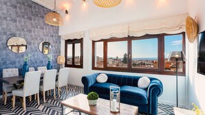 Ein modernes Wohnzimmer mit blauem Sofa, Esstisch und Stühlen, großen Fenstern mit Blick auf die Stadt und dekorativer Beleuchtung.