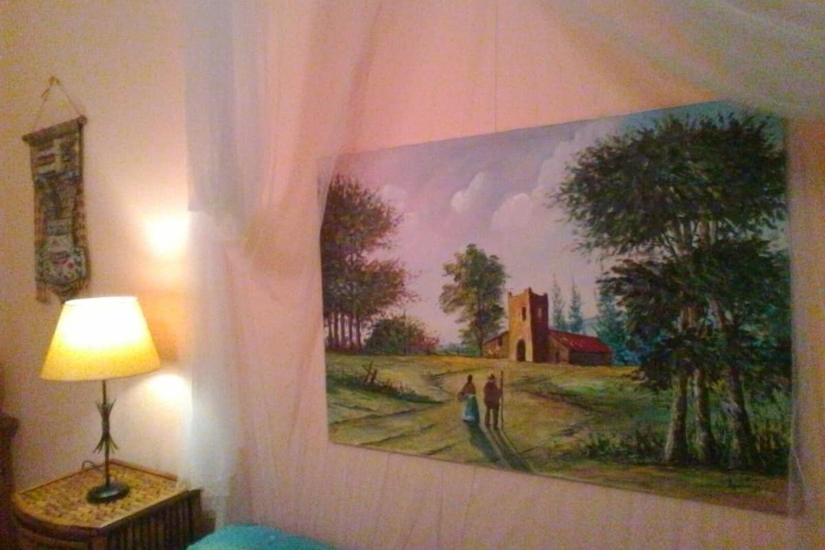 Una habitación con un cuadro de un paisaje rural, una lámpara en una mesita auxiliar y un tapiz en la pared.