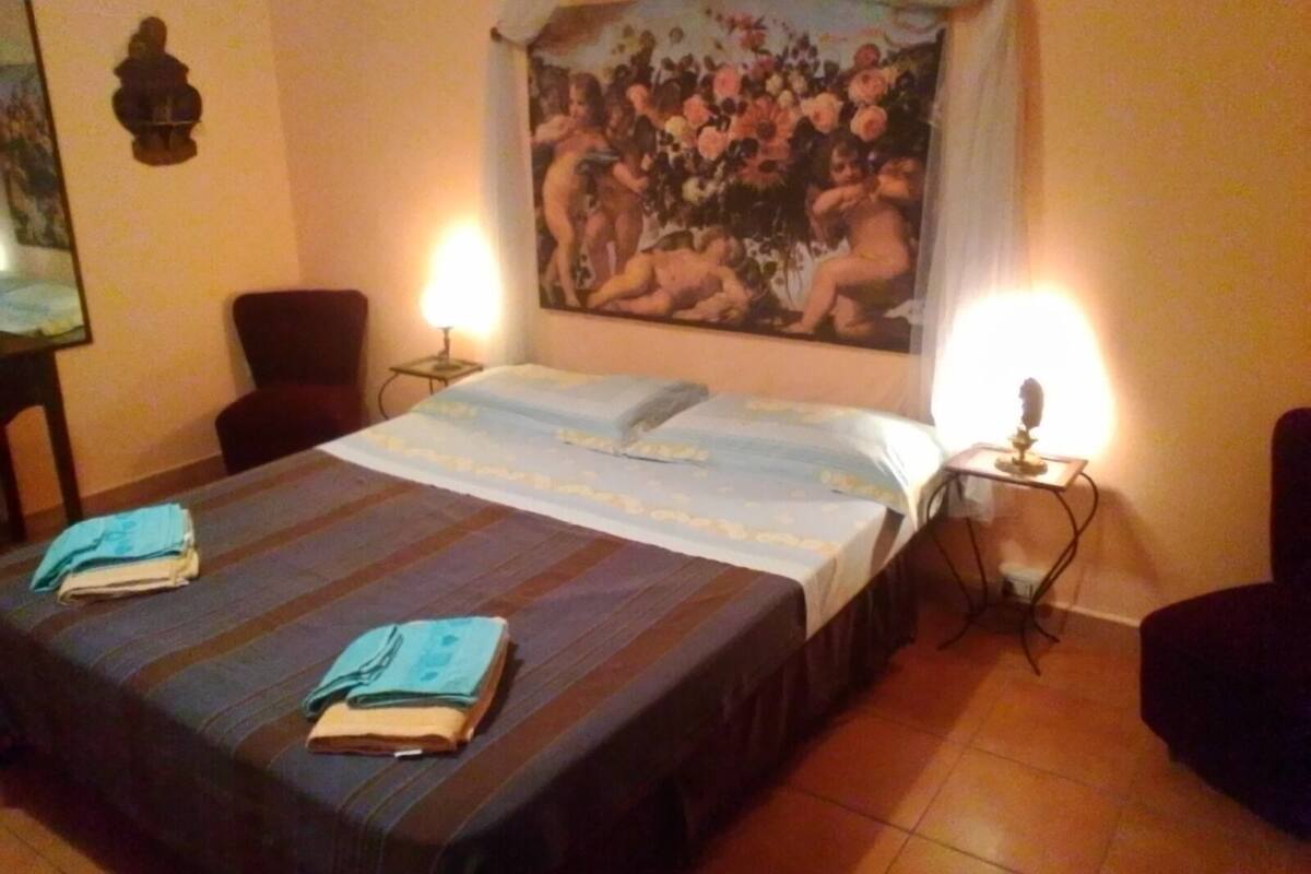 Habitación de hotel con cama, dos lámparas de noche, una silla, escritorio y un cuadro en la pared.
