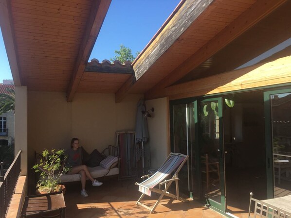 A covered outdoor area with a wooden ceiling, a cushioned seating area, a small table, and a potted plant.
