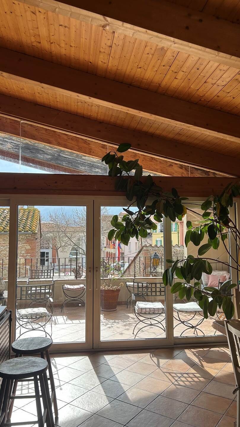 A covered patio with a view of a residential area, featuring a wooden ceiling, large sliding glass doors, and outdoor furniture.