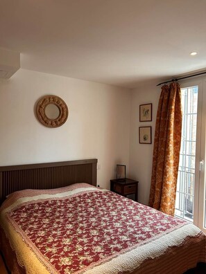 A bedroom with a large bed, a headboard, a small bedside table, a wall-mounted mirror, and a window with curtains.