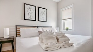Een netjes opgemaakt bed met gevouwen handdoeken, een nachtkastje met een lamp en twee ingelijste kunstwerken aan de muur.
