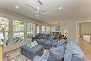 A spacious living room with a grey sectional sofa, a dining area with wicker chairs, and large windows with blinds.