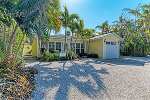 A yellow building with a white garage door, surrounded by palm trees and a gravel driveway.