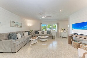 A modern living room with a grey sofa, two white coffee tables, a ceiling fan, and a large TV on a wooden stand. - Crescent Palms north end Ami, 2 King suites (Anna Maria)