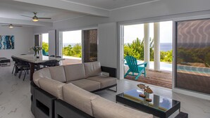 A modern living room with a large sofa, a dining table, and chairs. There are large windows with a view of the ocean.