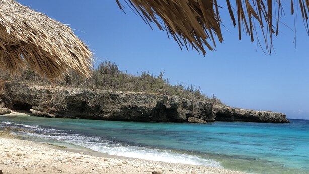 A beach with clear turquoise water, rocky outcrops, and a thatched structure.