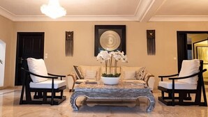 Ein geräumiger Wohnbereich mit einem beigen Sofa, zwei schwarzen Sesseln, einem Marmor-Couchtisch und einer Kronleuchter.